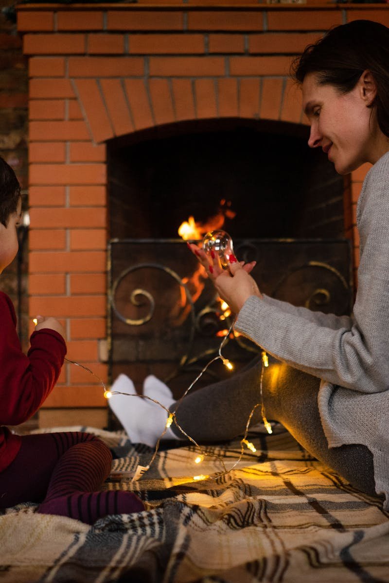 A mother and child share a joyful moment with lights by the fireplace indoors.